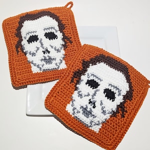 May include: Two square, orange crochet pot holders featuring a white mask with black eye holes and brown hair, resembling a horror movie character. Each pot holder has a small loop for hanging.