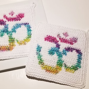 May include: Two white crochet potholders with a colorful Om symbol in rainbow hues. The potholders are square-shaped with a small loop for hanging. The Om symbol is a vibrant mix of pink, purple, blue, green, and yellow.