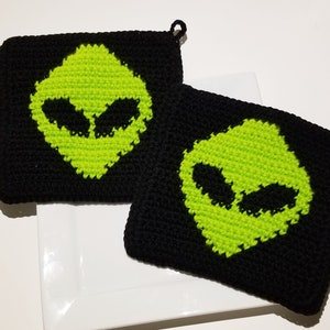 May include: Two black crocheted potholders with a neon green alien face design. Each potholder has a small loop for hanging. The potholders are square-shaped and rest on a white plate.
