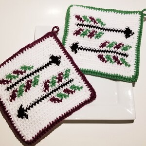Peut inclure: Deux dessous de plat crochetés à la main. L'un a une bordure bordeaux, l'autre une bordure verte. Les deux ont un centre blanc avec un motif de flèche noire, agrémenté de feuilles vertes et bordeaux. Chaque dessous de plat a une boucle pour suspendre.