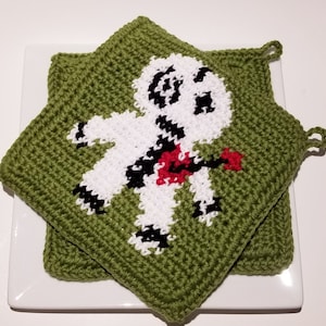 May include: Two green crocheted potholders with a white, black, and red design. The design features a figure with a red guitar. The potholders are square-shaped with a loop for hanging. The potholders are sitting on a white plate.