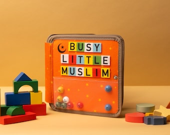 May include: An orange fabric activity book with the words "BUSY LITTLE MUSLIM" in colourful letter blocks. The book has a brown border and a variety of colourful wooden blocks and shapes are scattered around it.