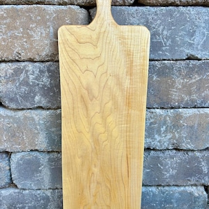 May include: A natural wood cutting board with a long, rectangular shape and a single handle. The board has a light brown color and a smooth, polished surface.