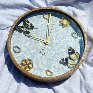 May include: A round, wooden clock with a blue and white patterned face. The clock has gold-colored hands and decorative accents, including butterflies and rings.