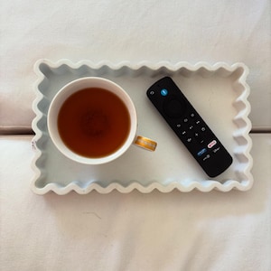 May include: A white, scalloped-edge tray with a cup of tea and a black remote control. The teacup has a gold band. The remote has several buttons and the Netflix and Disney+ logos.