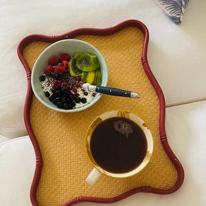 May include: A breakfast tray with a light brown woven surface and a red border holds a bowl of fruit and yogurt, a coffee cup, and a spoon. The bowl contains blueberries, raspberries, kiwi, and chia seeds. A pillow with a blue and white leaf pattern is in the background.