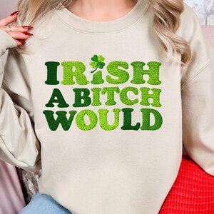 Op de afbeelding: Beige sweatshirt met de tekst "IRISH A BITCH WOULD" in groentinten. Het woord "IRISH" heeft een klaverblad-afbeelding. De sweatshirt is een casual stijl, perfect voor St. Patrick's Day of elke dag.