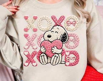 Könnte beinhalten: Beiges Sweatshirt mit Snoopy, der ein rosa Herz hält. Das Design umfasst gestrickte XOXO-Buchstaben in Rosa und Weiß sowie Herzakzente. Das Sweatshirt hat einen Rundhalsausschnitt und lange Ärmel.