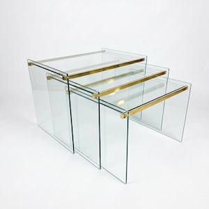 May include: A set of three nesting tables constructed from clear glass panels. Each table features a rectangular top supported by two side panels, with a horizontal gold-toned metal bar connecting the top to the sides at the front edge.