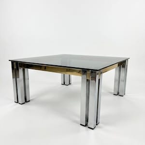 Può includere: Un tavolino quadrato con piano in vetro fumé scuro. La struttura è composta da quattro gambe, ciascuna formata da tre barre metalliche verticali cromate, collegate da una fascia metallica color oro sotto la superficie in vetro.