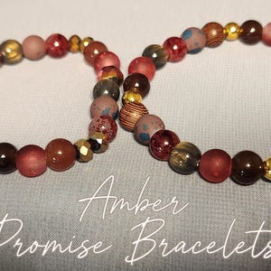 May include: Two beaded bracelets with the text "Amber Promise Bracelets". The bracelets feature a mix of amber, brown, red, and gold-colored beads. The beads are various sizes and shapes, creating a unique look.