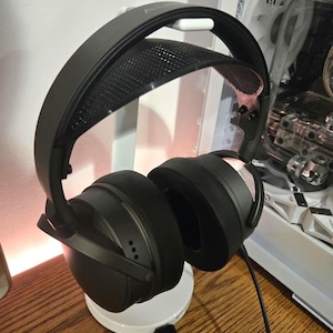 May include: Black over-ear headphones with a black headband and a white stand. The headphones are on a wooden surface.