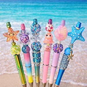 May include: A selection of decorative pens with sea-themed toppers, such as starfish and shells. The pens have metallic barrels in various colours, set against a beach backdrop with the ocean in the background.