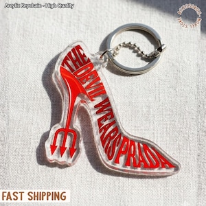 Puede incluir: Llavero acrílico transparente con forma de zapato de tacón alto con el texto rojo "THE DEVIL WEARS PRADA". El tacón tiene un diseño de tridente rojo. Incluye un llavero y una cadena plateados.