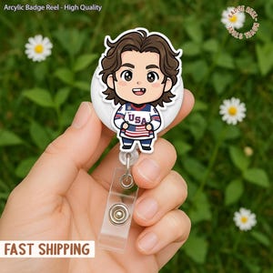 May include: A white badge reel with a cartoon illustration of a person wearing a USA hockey jersey. The badge reel has a clear plastic strap and a metal clip. The text "Arcylic Badge Reel - High Quality" is at the top.
