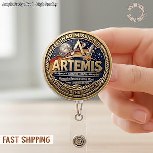 Op de afbeelding: Een goudkleurige Artemis badge reel met een gedetailleerd ontwerp met een raket, maan en sterren. De tekst bevat "LUNAR MISSION II", "ARTEMIS" en "RETURN TO THE MOON 2026". De badge reel is bevestigd aan een doorzichtige plastic houder.