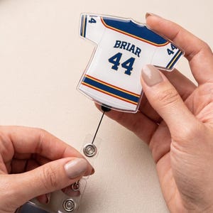 May include: A white and blue jersey-shaped badge reel with the name "Briar" and the number "44". The badge reel has a retractable cord and a clear plastic clip for attaching to an ID badge. The jersey has blue and gold stripes.