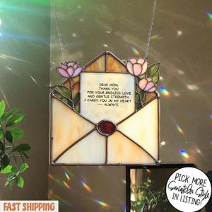 May include: A stained glass envelope-shaped suncatcher, featuring pink flowers and green leaves. The envelope displays the text "DEAR MOM, THANK YOU...I CARRY YOU IN MY HEART - ALWAYS." A red gem is in the center.