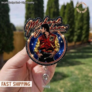 May include: An acrylic badge reel featuring a portrait of Michael Jackson in a red and gold jacket. The design includes the name "Michael Jackson" in a glittery font. The reel has a clear plastic clip and the text "Arcylic Badge Reel - High Quality".