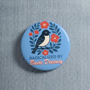 Może przedstawiać: Okrągła, jasnoniebieska przypinka z czarno-białym ptakiem siedzącym na gałęzi, otoczonym czerwonymi kwiatami i zielonymi liśćmi. Tekst "RADICALIZED BY Basic Decency" jest napisany na czerwono i biało.