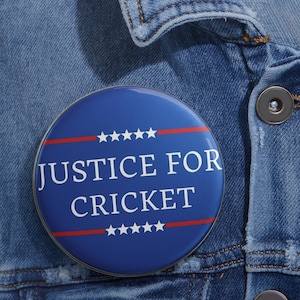 May include: A round blue button with the text "JUSTICE FOR CRICKET" in white, with red stripes and white stars. The button is pinned to a denim jacket.