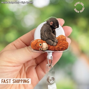 Puede incluir: Un carrete de insignia con una cubierta de plástico transparente que presenta una ilustración detallada de un mono acostado encima de un orangután. El carrete tiene una base blanca y un cordón retráctil. El texto "Arcylic Badge Reel - High Quality" está en la parte superior.