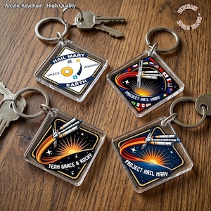 Puede incluir: Llaveros de acrílico transparente con diseños espaciales, cohetes, estrellas y los textos "HAIL MARY", "TEAM GRACE & ROCKY" y "EARTH". Cada llavero tiene un anillo plateado. Se muestran sobre una superficie de madera.