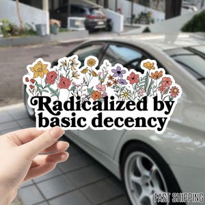 May include: A white sticker with a floral design and the text "Radicalized by basic decency." The sticker features a variety of colorful flowers, including yellow, red, orange, and purple, with green leaves. The sticker is held in front of a car.