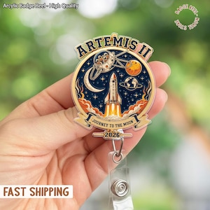 Op de afbeelding: Een acryl badge reel met een ruimtevaartthema. De reel toont de tekst "ARTEMIS II" en "JOURNEY TO THE MOON 2026", met illustraties van een raket, planeten en een ruimteschip. Het heeft een transparant intrekbaar koord en een metalen clip.