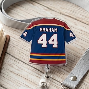 May include: A clear plastic badge reel featuring a miniature hockey jersey design. The jersey is navy blue with red and gold accents, displaying the name "GRAHAM" and the number "44". A gray lanyard and ID holder are also visible.