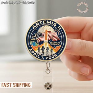 Op de afbeelding: Een ronde Artemis II badge reel met een blauw en goud ontwerp. De badge toont een lancering van een raket, Amerikaanse vlaggen en de tekst "APRIL 1, 2026". De tekst "FOR ALL HUMANITY" en "RETURN TO THE MOON" zijn ook aanwezig.