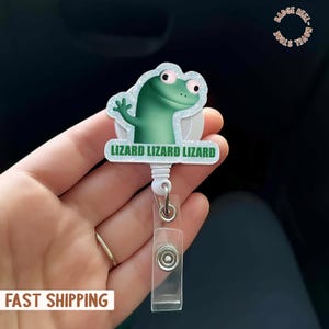 Puede incluir: Un portatarjetas con un diseño de lagarto verde y el texto "LIZARD LIZARD LIZARD" en verde. El portatarjetas tiene una correa de plástico transparente y un clip de metal plateado. El lagarto tiene ojos rosas y una expresión amigable.