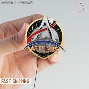 Op de afbeelding: Een ronde Artemis badge reel. Het ontwerp omvat een gouden rand, een zwarte achtergrond met sterren en een centraal ontwerp met een maan, een rode en witte boog en het woord "ARTEMIS". De tekst "Arcylic Badge Reel - High Quality" staat bovenaan.
