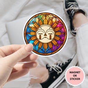 Puede incluir: Una pegatina o imán redondo con un diseño de vidriera de sol. El sol tiene una cara con un diseño estilizado. La pegatina la sostiene una persona. El texto "MAGNET OR STICKER" está en una pegatina rosa.
