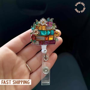 May include: A badge reel featuring a stack of colorful books adorned with flowers. The books are in shades of orange, teal, yellow, and purple. The reel has a clear plastic ID holder and a white retractable cord. The text "FAST SHIPPING" is visible.