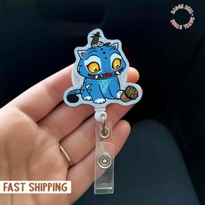 May include: A blue cat-like character badge reel with a glittery border. The cartoon cat has yellow eyes, a small hat, and a small pot of flowers. The badge reel has a clear plastic ID holder and a metal clip.