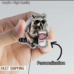 May include: A silver glitter-edged acrylic pin featuring a raccoon wearing sunglasses and drinking from a Dr. Pepper Diet can. The pin has the text "Arcylic - High Quality" at the top and "Personalization" at the bottom.