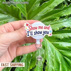 May include: A light blue retractable badge reel with the text "Show me and I'll show you" in red and blue. The badge reel has a clear plastic clip and a metal clasp. The background is a large green leaf.