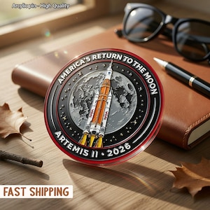 Op de afbeelding: Een ronde, hoogwaardige acryl pin met een rode rand. De pin toont een raket die vanaf de maan lanceert, met de tekst "AMERICA'S RETURN TO THE MOON" en "ARTEMIS II 2026". De pin ligt op een houten oppervlak.
