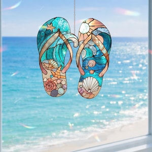 May include: A stained glass artwork showing two flip-flops with a beach design. The flip-flops feature ocean waves, seashells, and a starfish in blue, teal, and beige. The artwork is suspended by a chain against a blurred ocean backdrop.