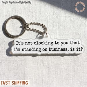 May include: Clear acrylic keychain with a silver keyring and chain. The keychain has the phrase "It's not clocking to you that I'm standing on business, is it?" printed in black. The keychain is a novelty item.