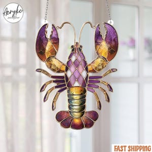 Op de afbeelding: Een gebrandschilderde glazen zonnevanger in de vorm van een kreeft. De kreeft heeft tinten paars, goud en wit, met een gedetailleerd patroon. Het hangt aan een ketting, perfect voor raam- of wanddecoratie.