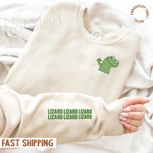 May include: A cream-coloured sweatshirt with a green cartoon lizard embroidered on the chest. The sleeve features the text "LIZARD LIZARD LIZARD LIZARD LIZARD LIZARD" in green. The words "EMBROIDERY SHIRT - NOVEL" are printed in a circle on the upper right.