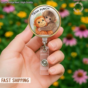 May include: A badge reel with a gold rim and a clear plastic strap. The reel features a cartoon image of two monkeys hugging, with the text "TEAM PUNCH" in a circular design. The background is a blurred image of flowers.