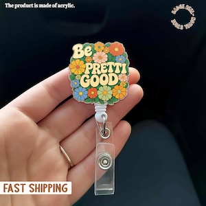 Pode incluir: Um porta-crachá floral com a frase "Be Pretti Good" rodeada por flores coloridas. O porta-crachá é feito de acrílico e possui um clipe de plástico transparente e um cordão retrátil.