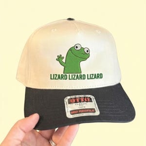 Könnte beinhalten: Baseballcap mit cremefarbener Krone und schwarzem Schirm. Die Kappe zeigt eine grüne Eidechsen-Grafik und den Text "LIZARD LIZARD LIZARD" in Grün. Die Kappe ist eine klassische Passform, OSFM und Mid-Profile.
