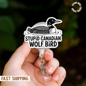 May include: A white and black badge reel with a cartoon loon illustration. The text "STUPID CANADIAN WOLF BIRD" is printed below the loon. The reel has a clear plastic strap and a metal clip.
