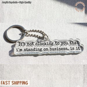 May include: A clear acrylic keychain with a silver keyring and chain. The keychain has the phrase "It's not clocking to you that I'm standing on business, is it?" printed in black. The keychain is a novelty item.