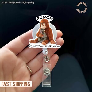 May include: A badge reel featuring a cartoon orangutan and baby monkey. The orangutan is brown, and the baby monkey is gray. The text "PUNCH" is above the orangutan, and "MY EMOTIONAL SUPPORT ANIMAL" is below. The reel is white with a clear plastic ID holder.