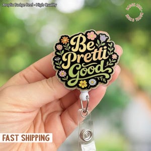 Puede incluir: Un carrete de insignia redondo con las palabras "Be Pretti Good" en una fuente caprichosa, rodeado de detalles florales y de hojas. El carrete de la insignia es negro con detalles amarillos, verdes y rosas. Tiene una correa de plástico transparente y un clip metálico.